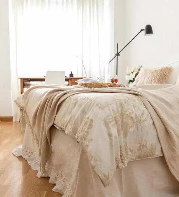 Dormitorio acogedor con cama vestida en tonos beige y blanco, con un escritorio de madera y luz natural en proyecto de Home Staging en Puig Santa María, Valencia.